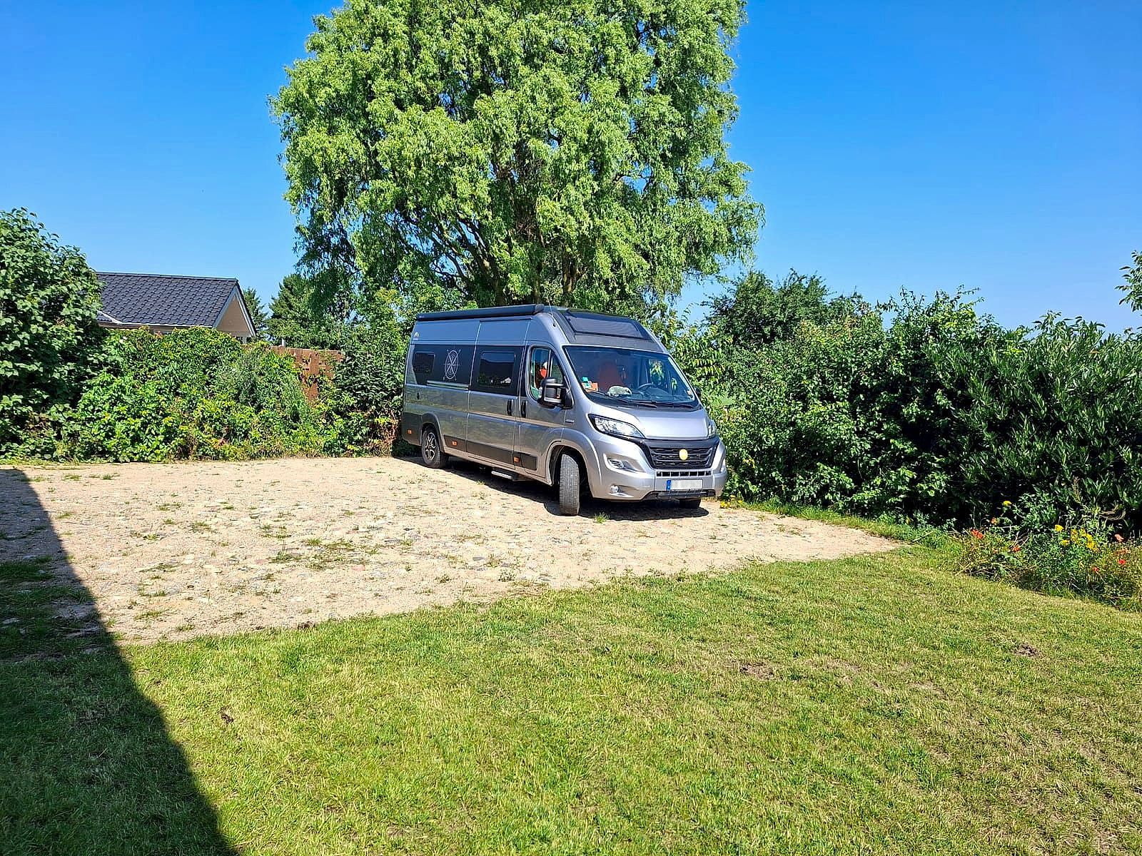 Motorhome on the lightly paved pitch right next to HAUS HOPPE gallery, with a large tree and open countryside behind