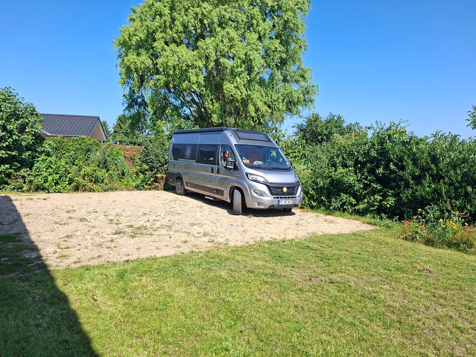 Motorhome on the lightly paved pitch right next to HAUS HOPPE gallery, with a large tree and open countryside behind