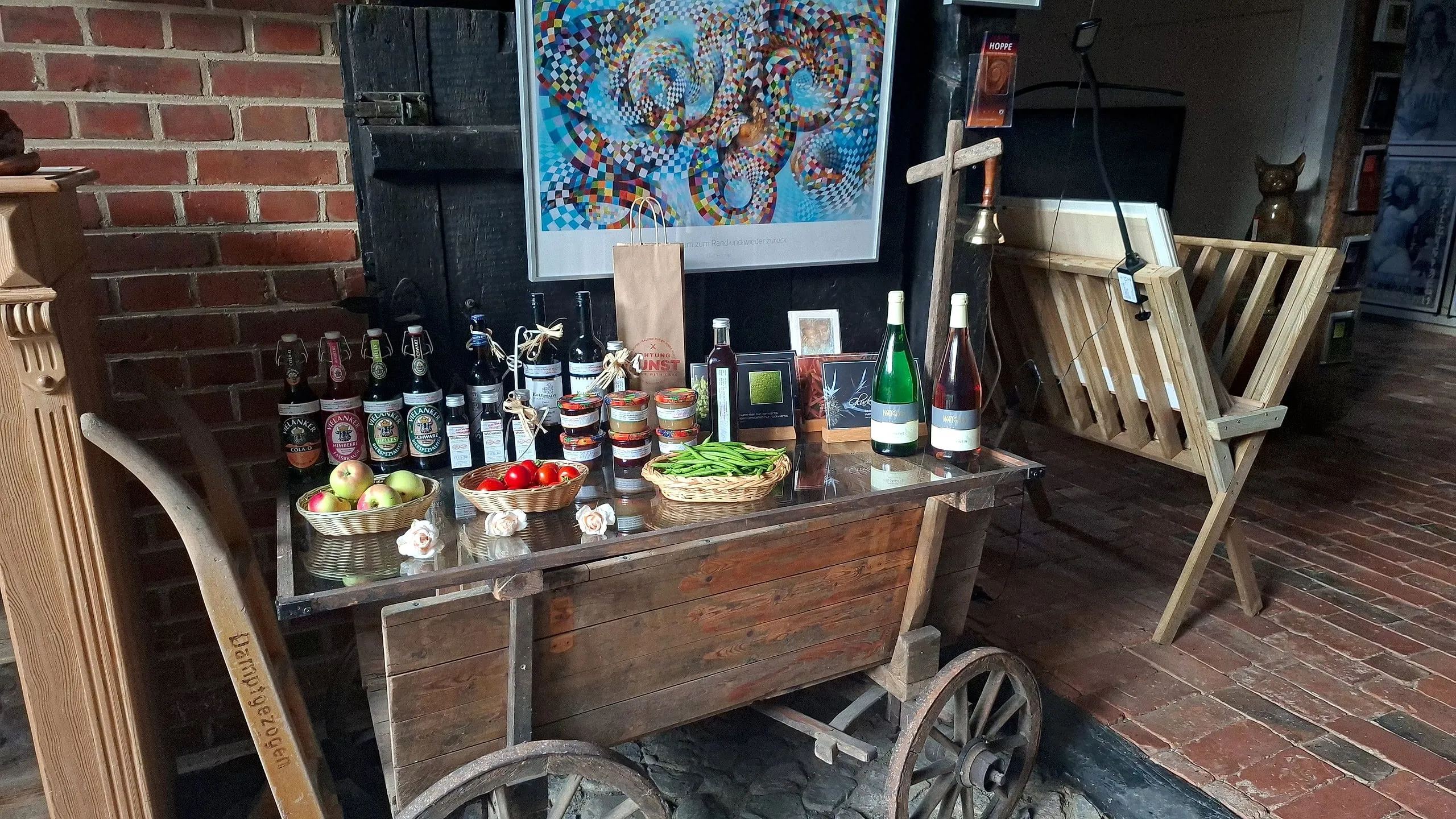 Old market cart with a glass top in the farm shop, holding beer, jam, oils and baskets of apples, tomatoes and beans, with a large colourful artwork on the brick wall behind